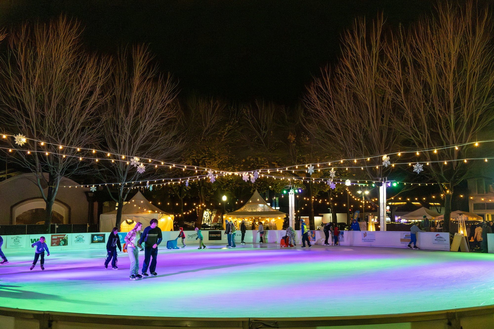 A vibrant outdoor ice skating rink at night, with festive lights, people skating, and decorated trees in the background.