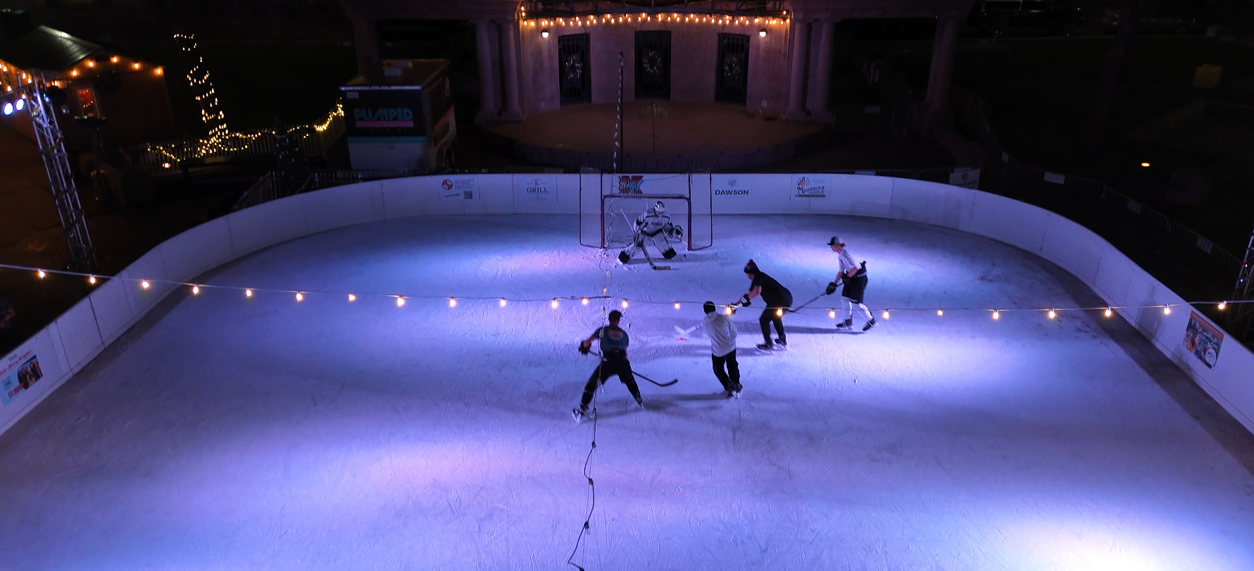 Nighttime hockey game on an outdoor rink with festive lights and multiple players in action.