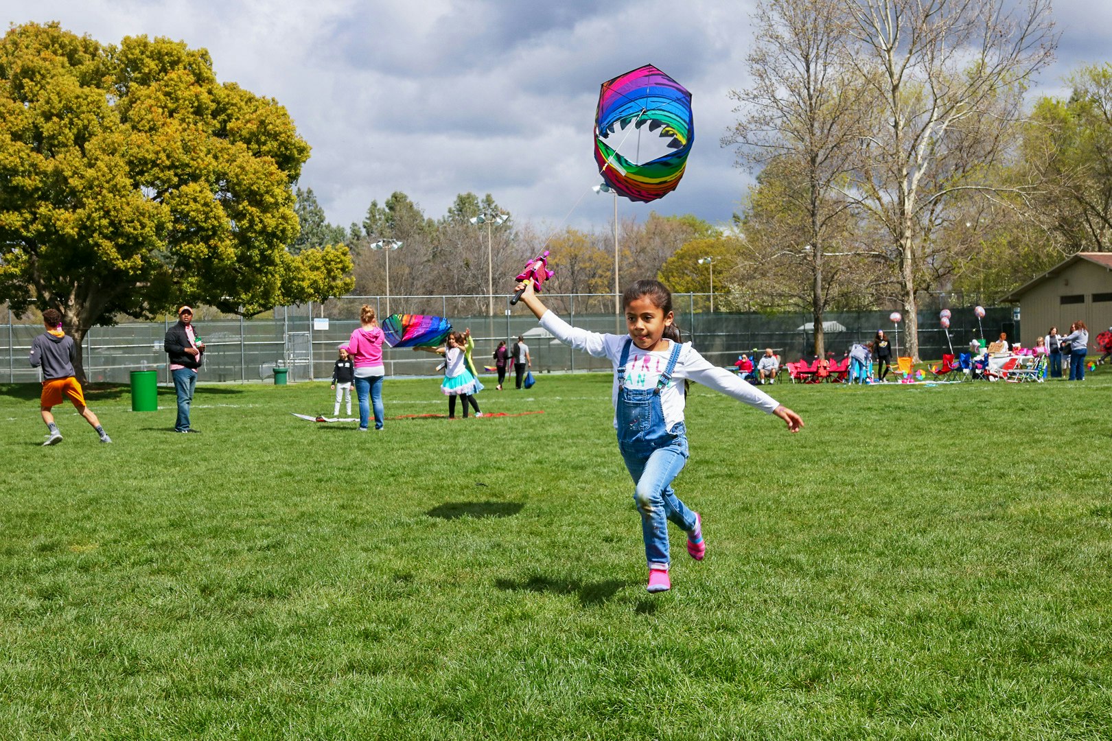 A girl running with a colorful kite in a park with others enjoying the outdoors.