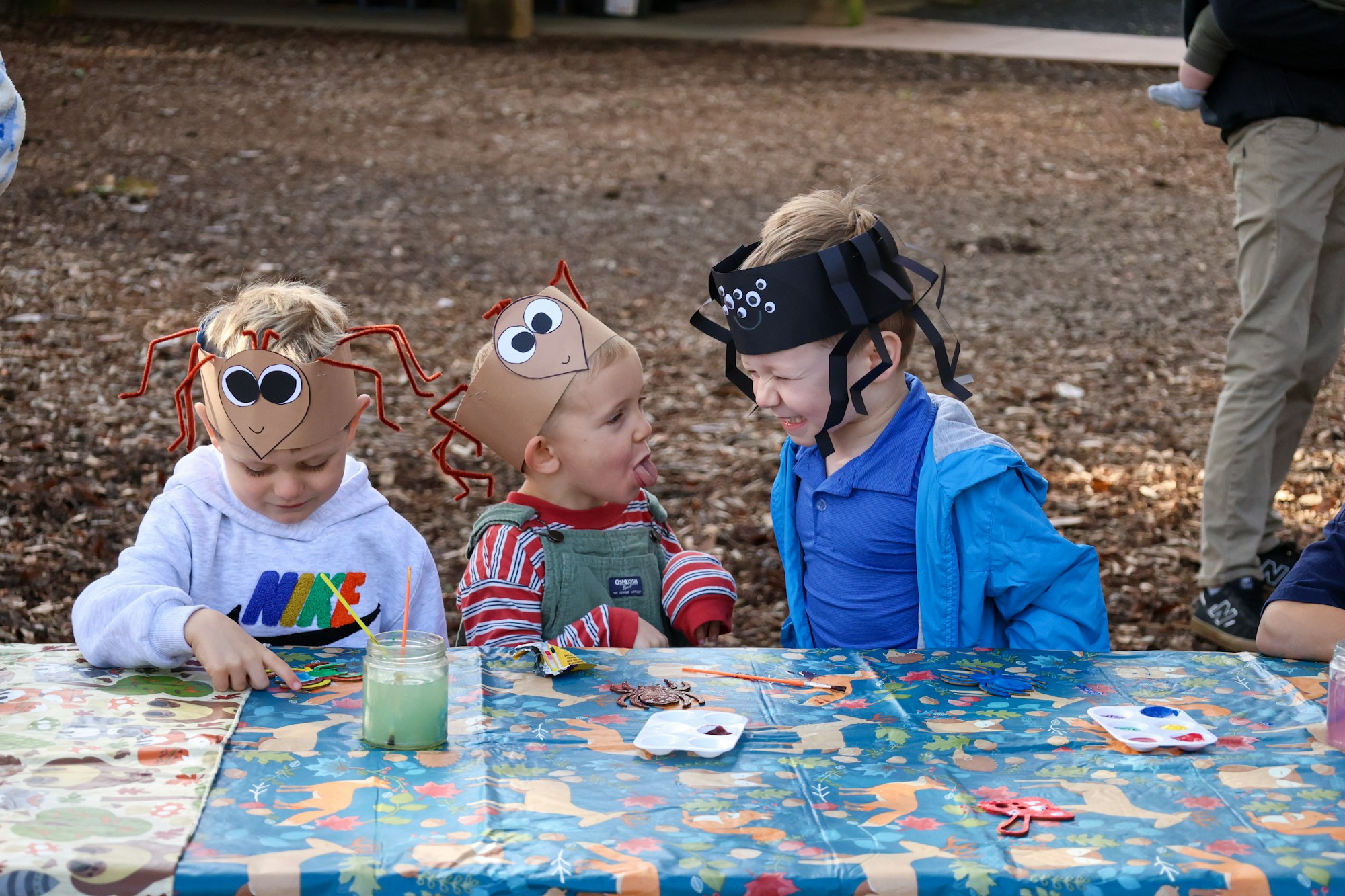 Three kids wearing funny spider-themed hats are sitting at a table, engaging in activities and having fun outdoors.