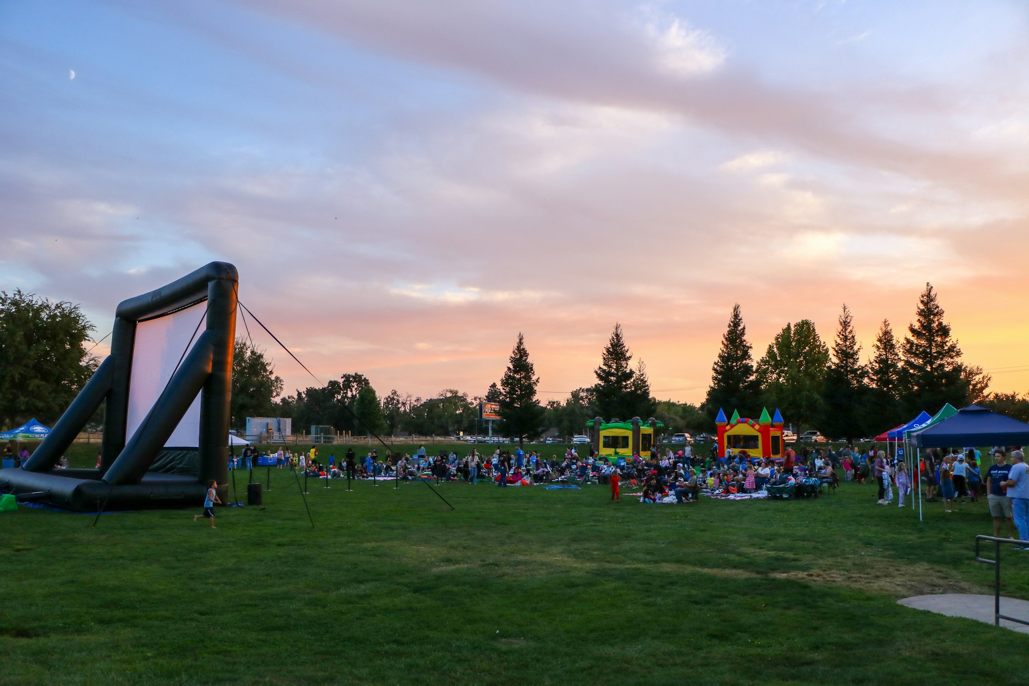Outdoor event with an inflatable screen, people on the grass, bounce houses, and a sunset sky.