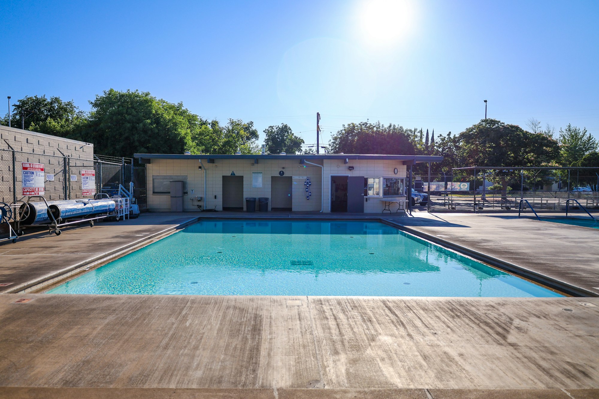 Outdoor swimming pool with concrete deck, building, and trees in the background under a bright, clear sky.