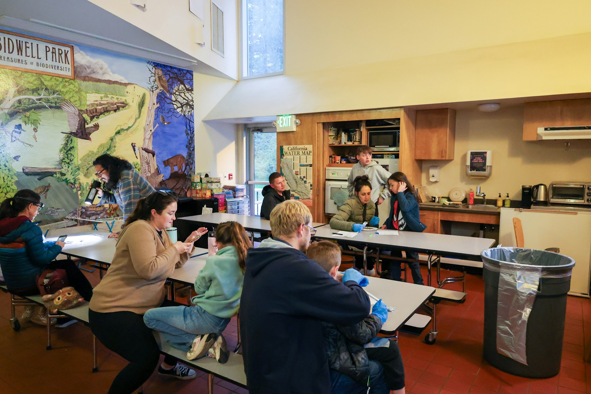 People seated in a classroom with a mural on the wall, engaged in various activities, including using phones and writing.