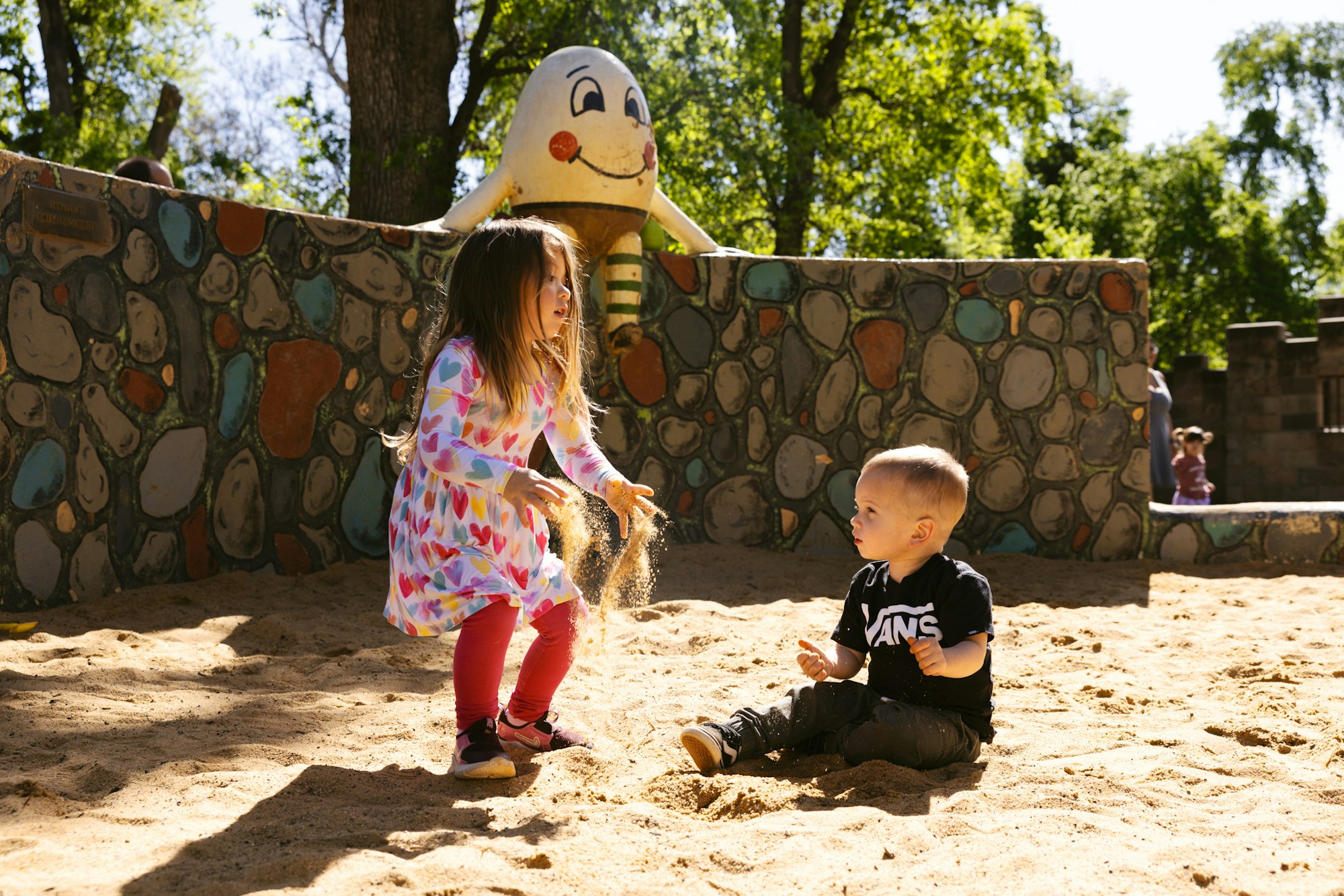 A girl in a heart-patterned dress plays in a sandbox while a boy in a black shirt observes, with a colorful wall in the background.
