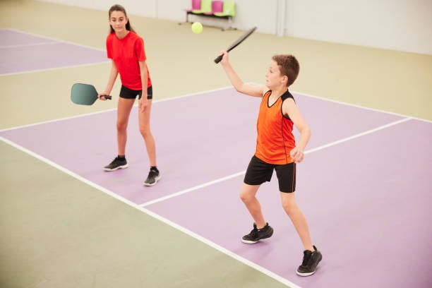 A boy and a girl are playing a racket sport indoors, with the boy preparing to hit a ball while the girl stands ready.