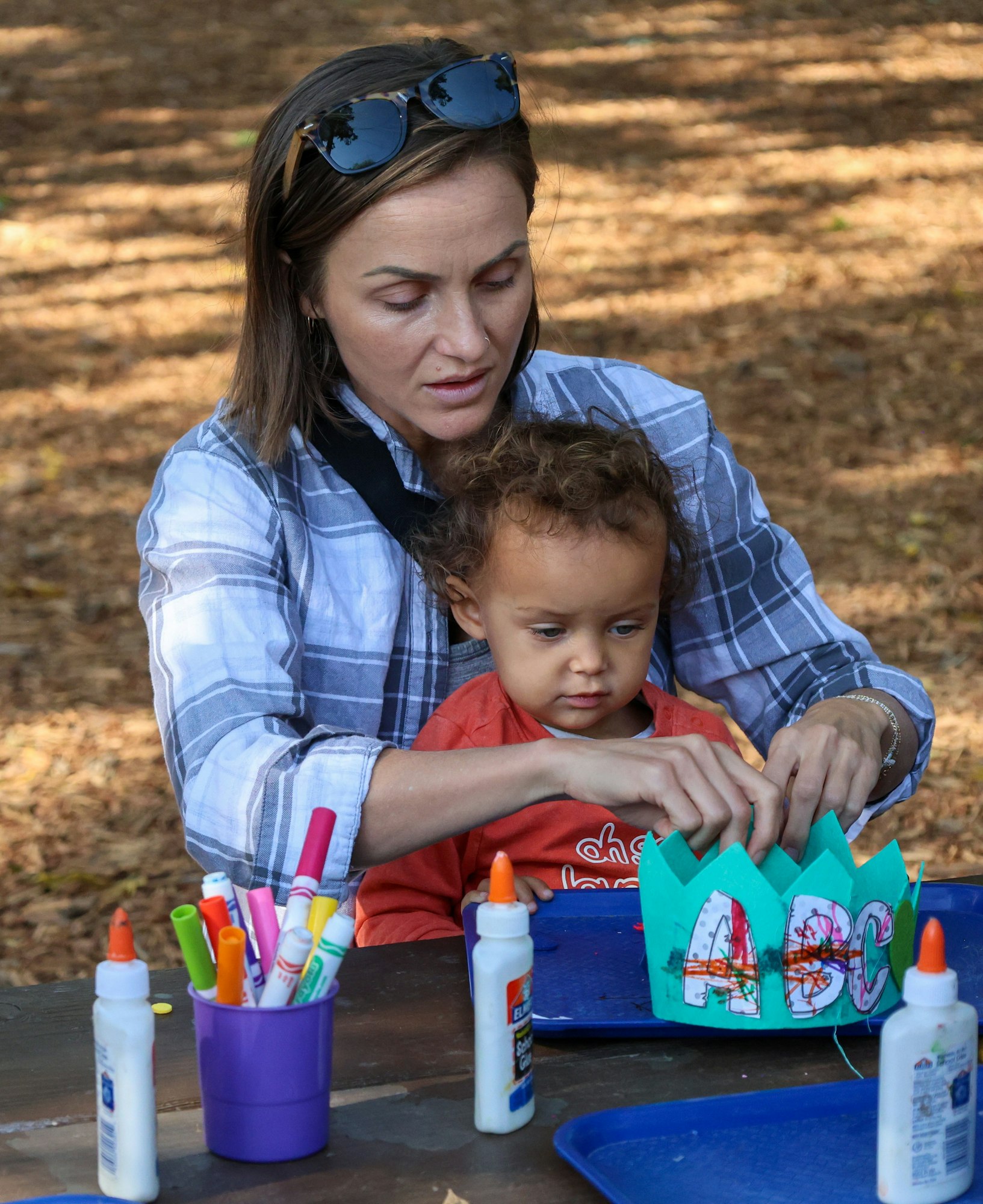 A woman helps a child craft a decorated crown with "ABC" using glue and markers at a outdoor table.