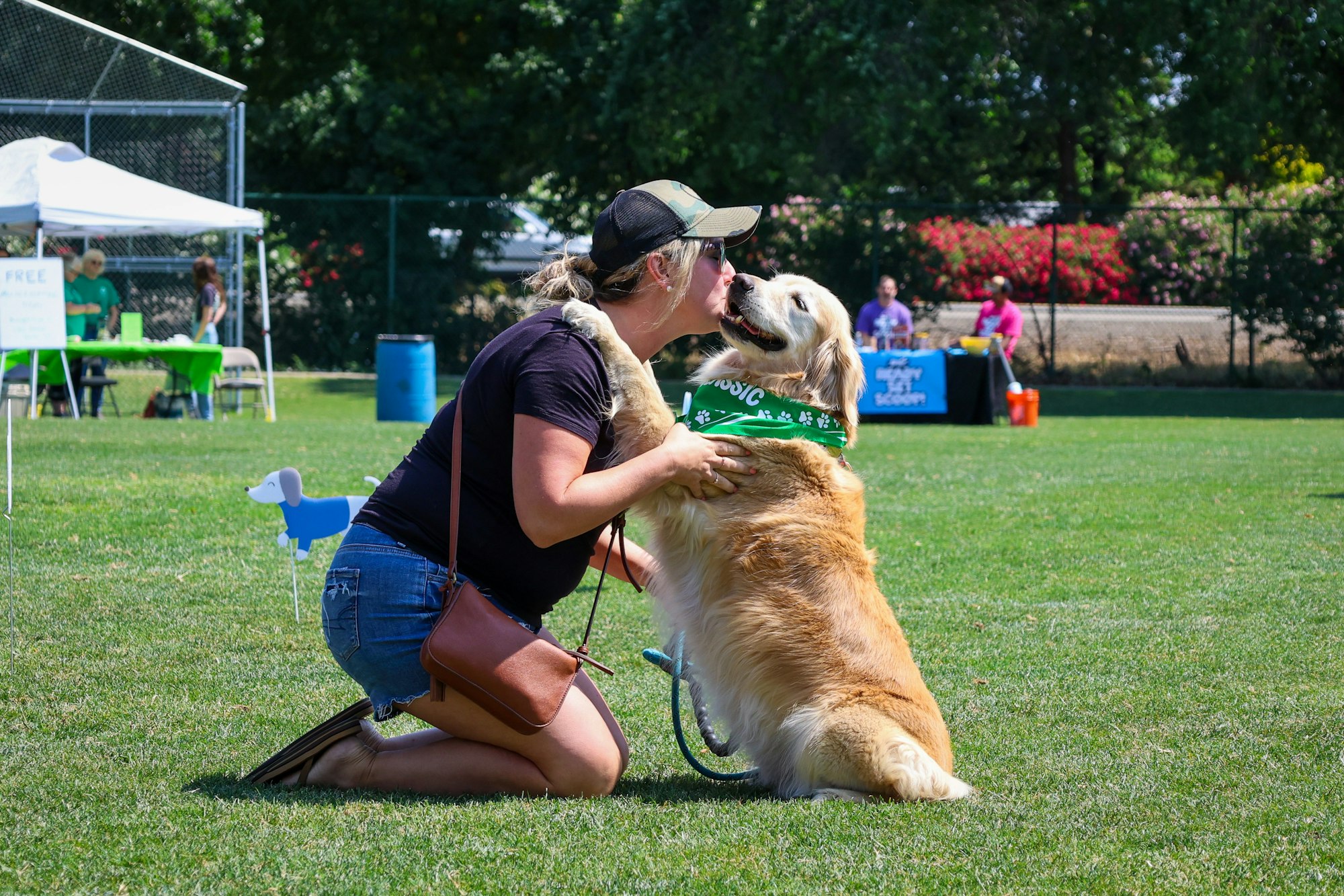 A person kneels to kiss a golden retriever wearing a green bandana in a park setting with tents and people in the background.