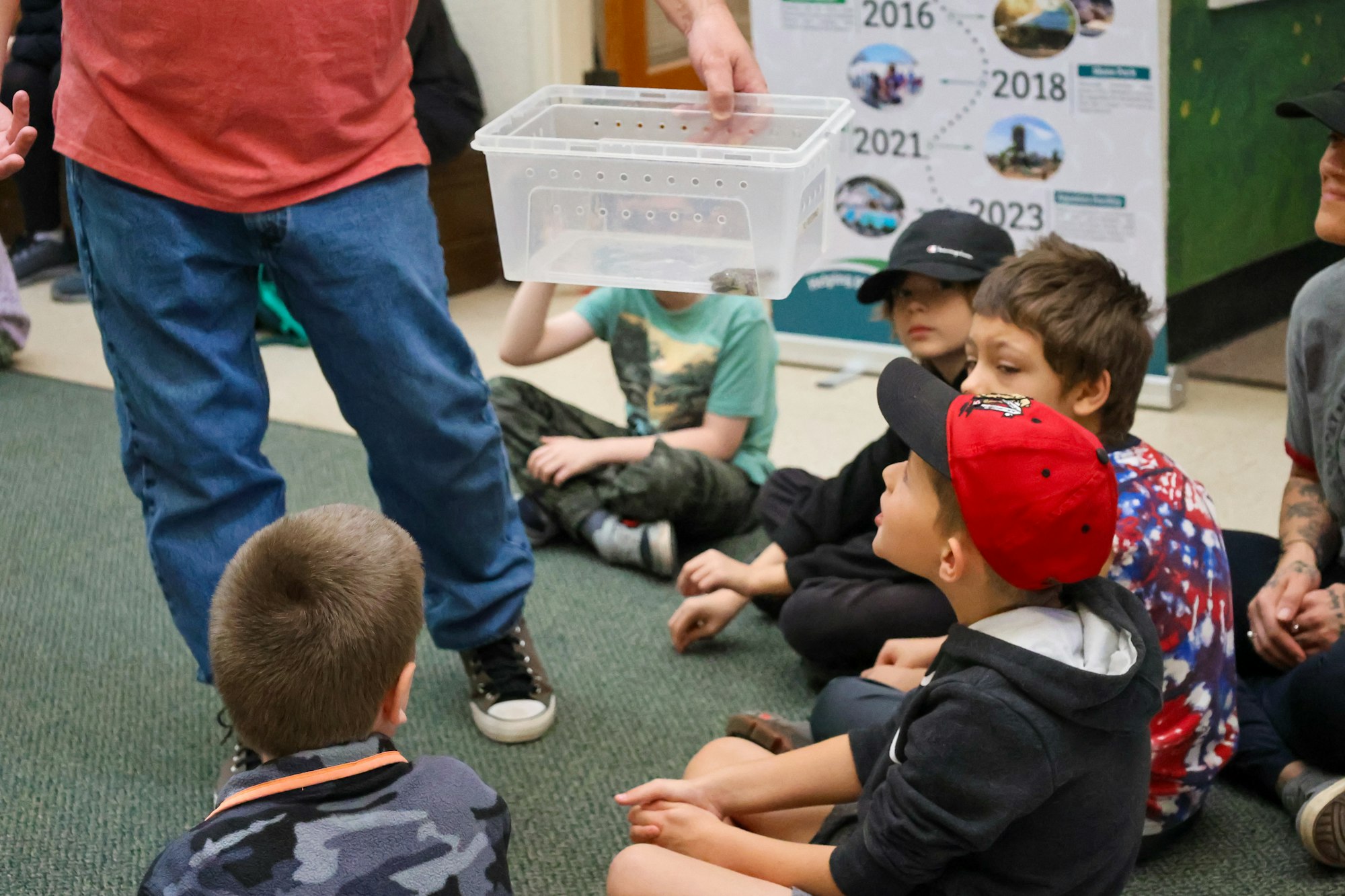 Children sitting on the floor watching a person holding a clear container, possibly for an educational demonstration.