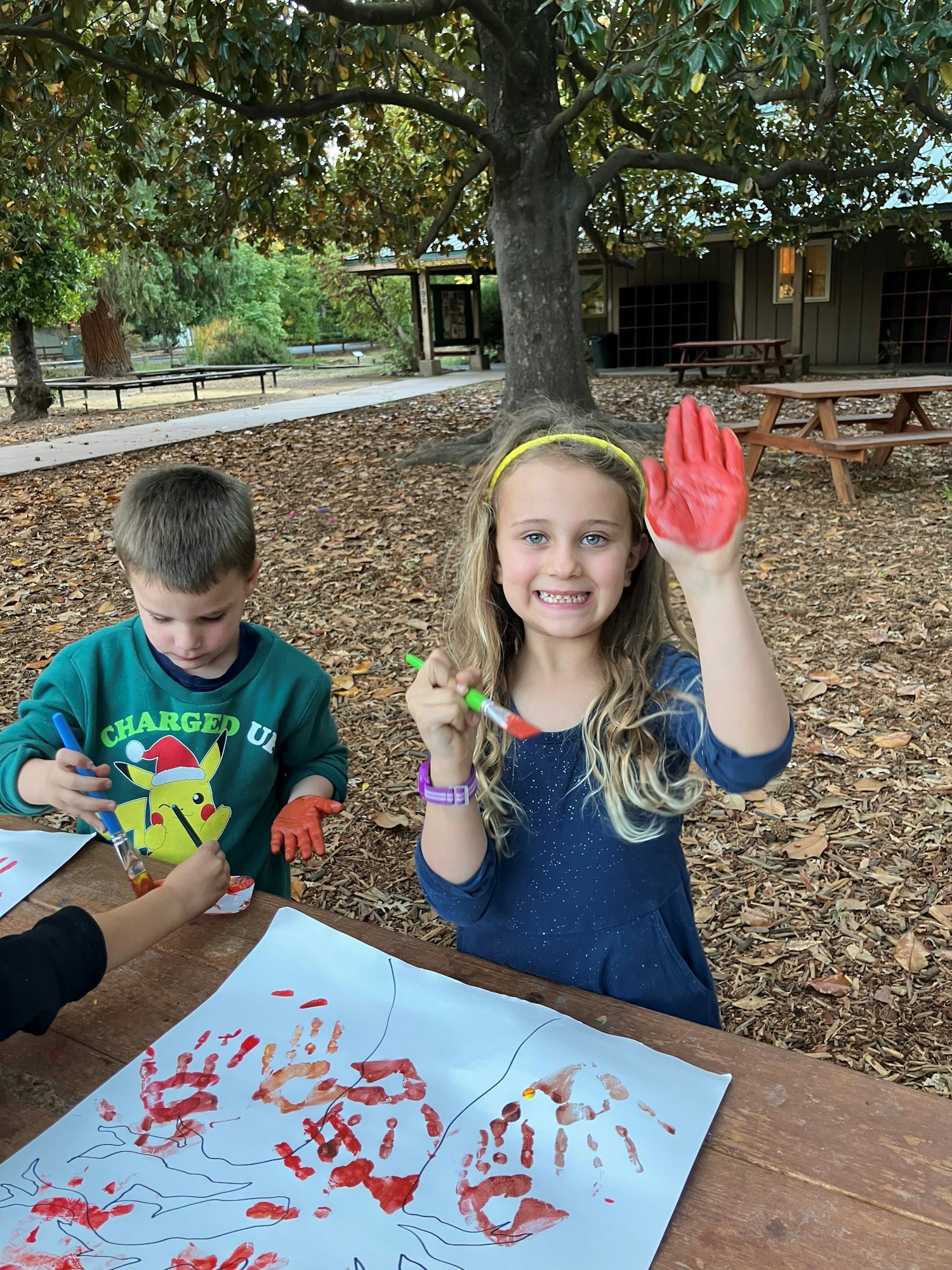Kids doing handprint painting outdoors under a tree.