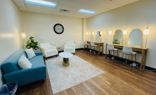 A cozy lounge with a sofa, chairs, tables, mirrors, and bright lighting. Decor includes a clock, plants, and a rug on wooden flooring.