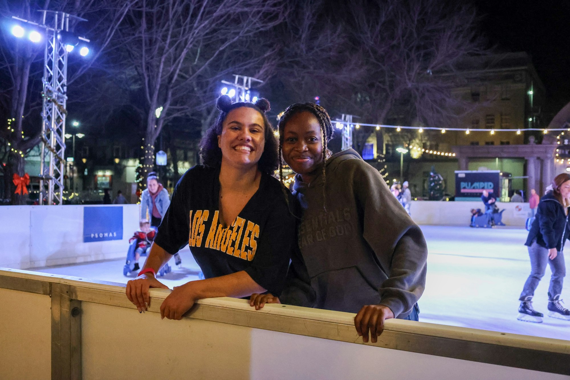 Two people smiling by an outdoor ice rink, with lights and skaters in the background.