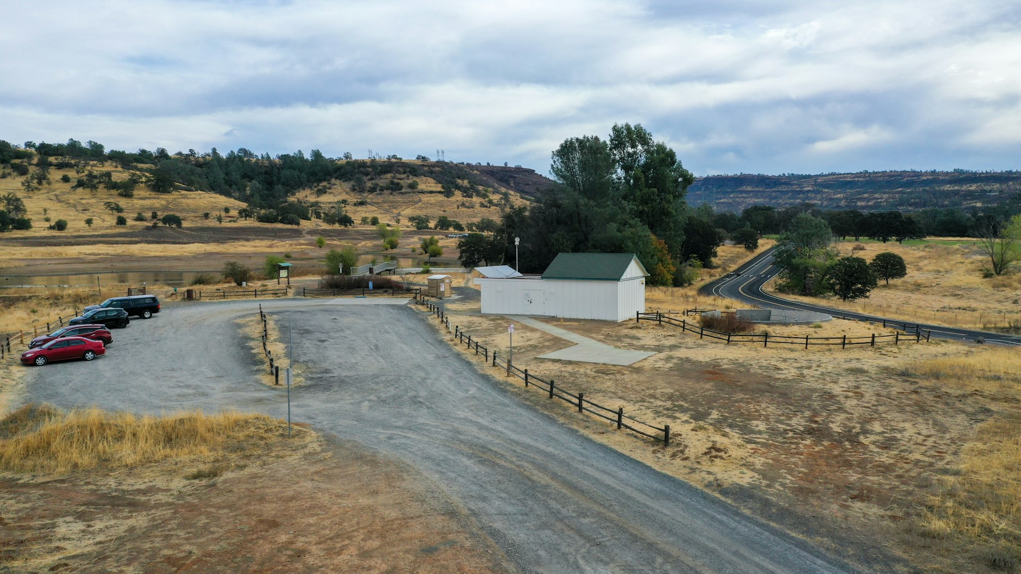 Rural landscape with a small building, parking area, road, and grassy hills under a cloudy sky.