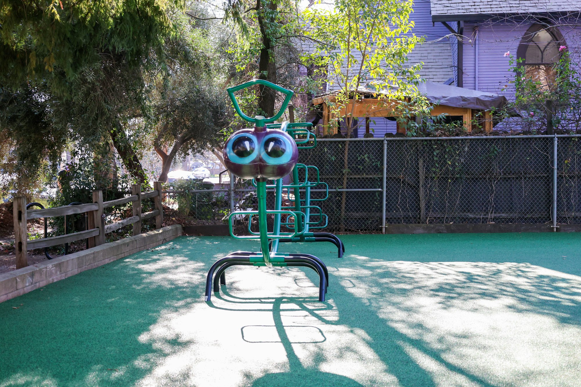 A playground feature resembling a bug with large eyes, set on green turf, surrounded by trees and a fence.