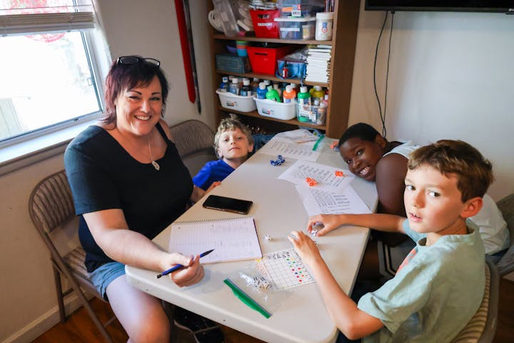 A woman and three children sitting at a table with paper, dice, and art supplies, smiling and engaging in an activity.