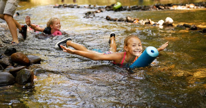 Two children are playing and floating in a shallow stream, enjoying the water with pool noodles. Bright and cheerful atmosphere.