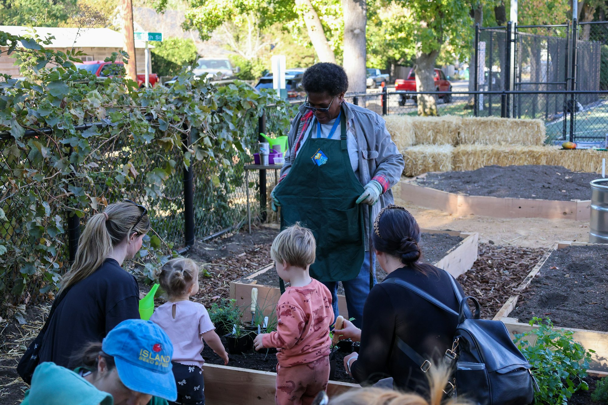 A group of people, including children, are gardening together outside near raised beds and hay bales.