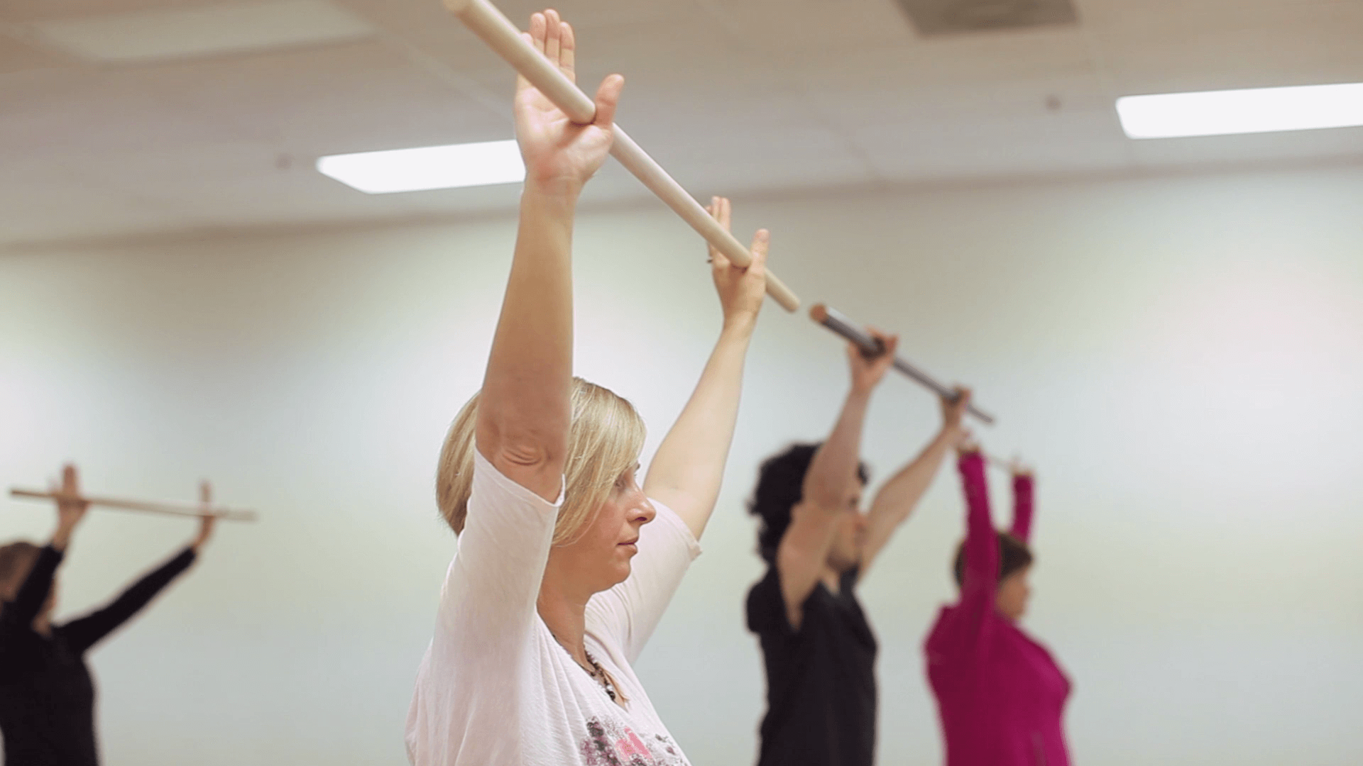 People in a fitness class holding poles overhead, exercising in a line.