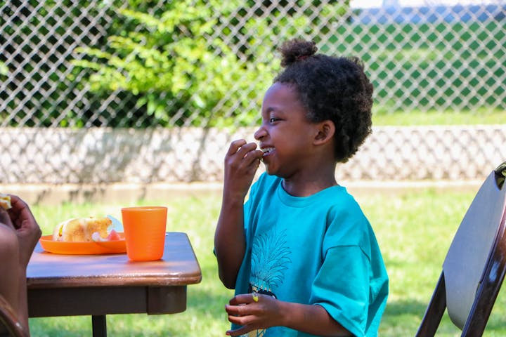 A child eating at a table outdoors with a fence and greenery in the background.