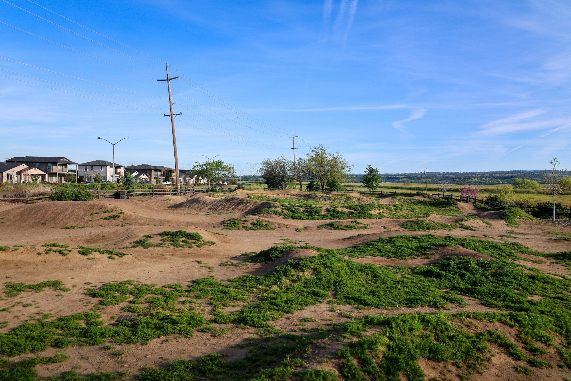 A BMX or mountain bike track with dirt jumps, set near a housing area under a clear sky.