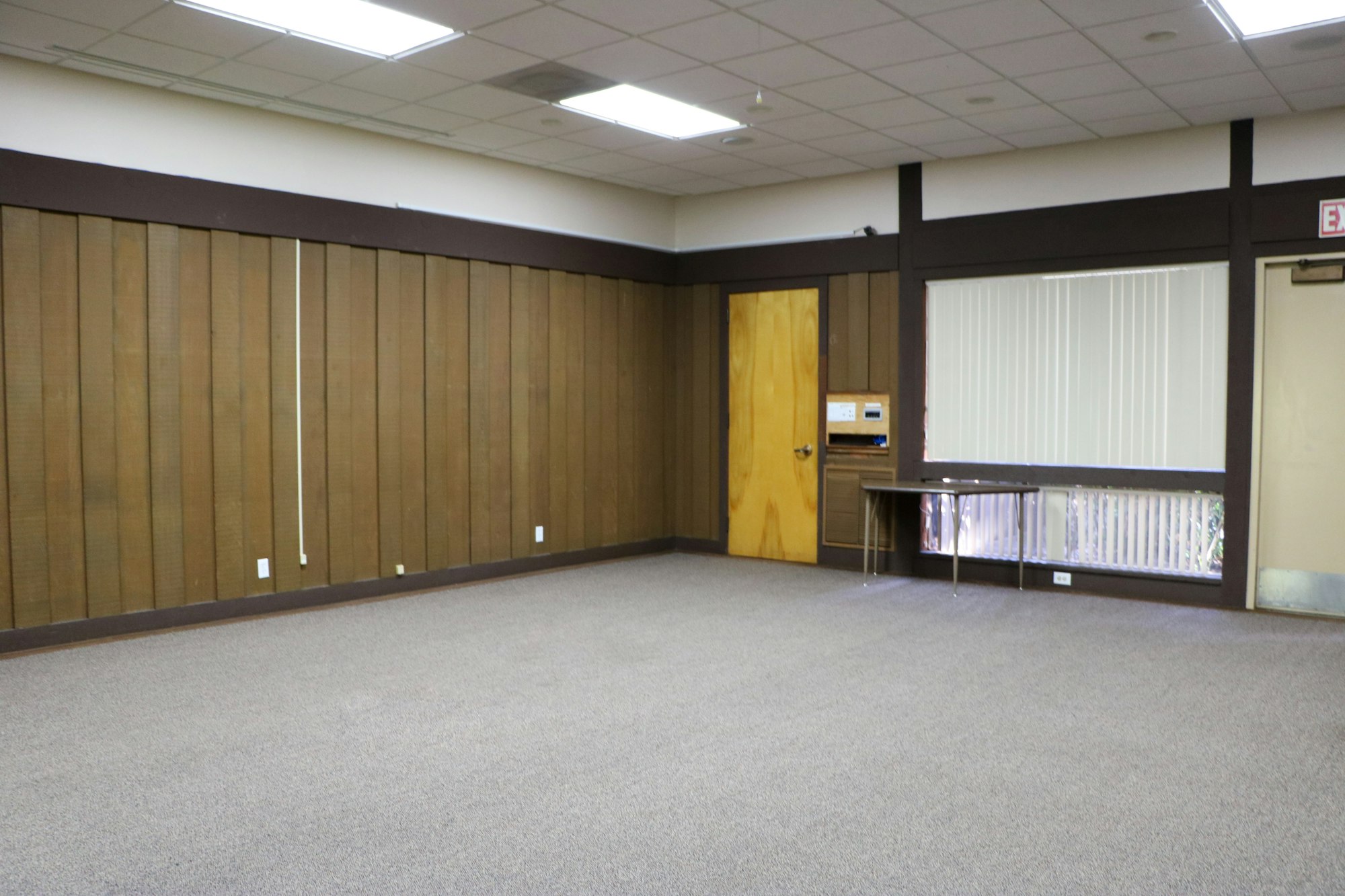 An empty room with wood-panel walls, a door, blinds-covered window, and gray carpet.