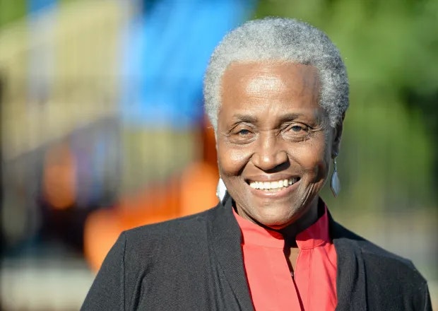 An older person smiling, wearing a red shirt and dark jacket, with a blurred colorful background.