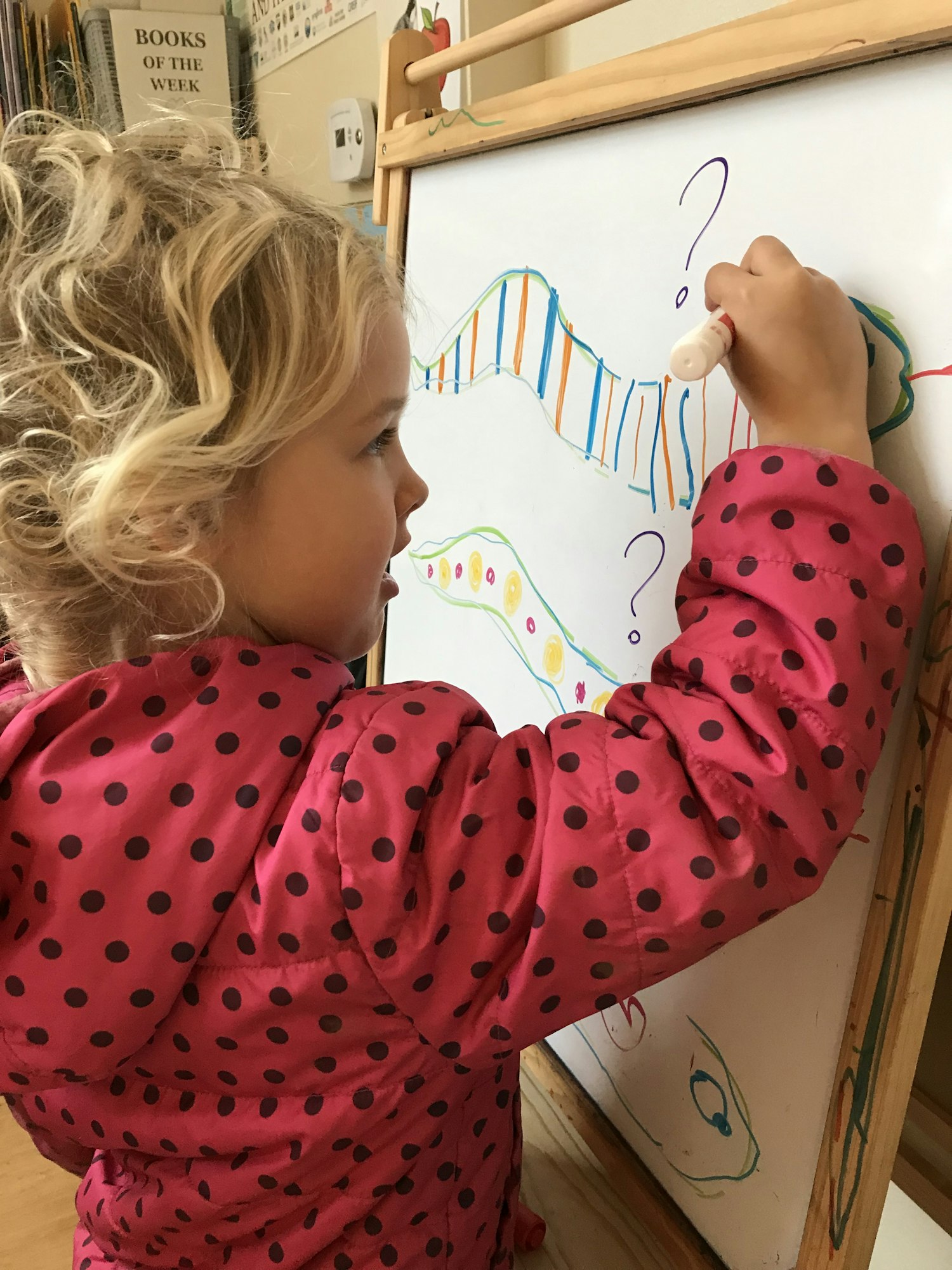 Child drawing with markers on a whiteboard, wearing a pink polka dot jacket.