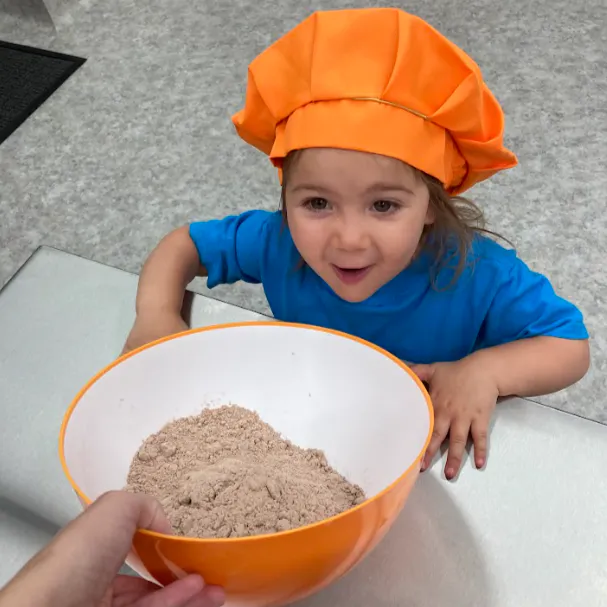A young child in an orange chef hat is excitedly looking at a bowl of flour or dry mix being presented to them.