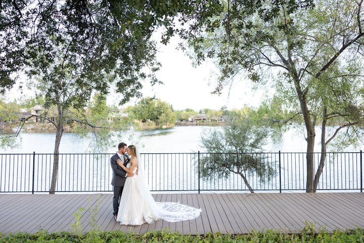 Bride and groom embracing on a wooden deck by a lake, surrounded by trees.