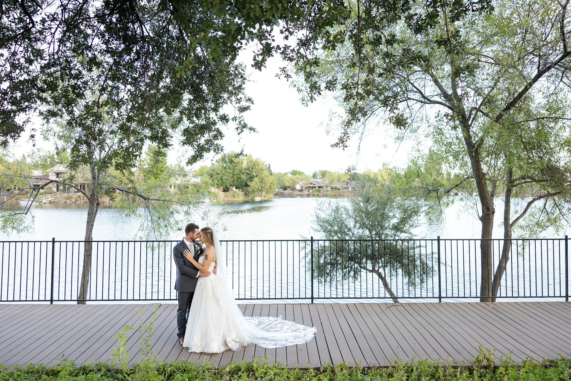 Bride and groom embracing on a wooden deck by a lake, surrounded by trees.