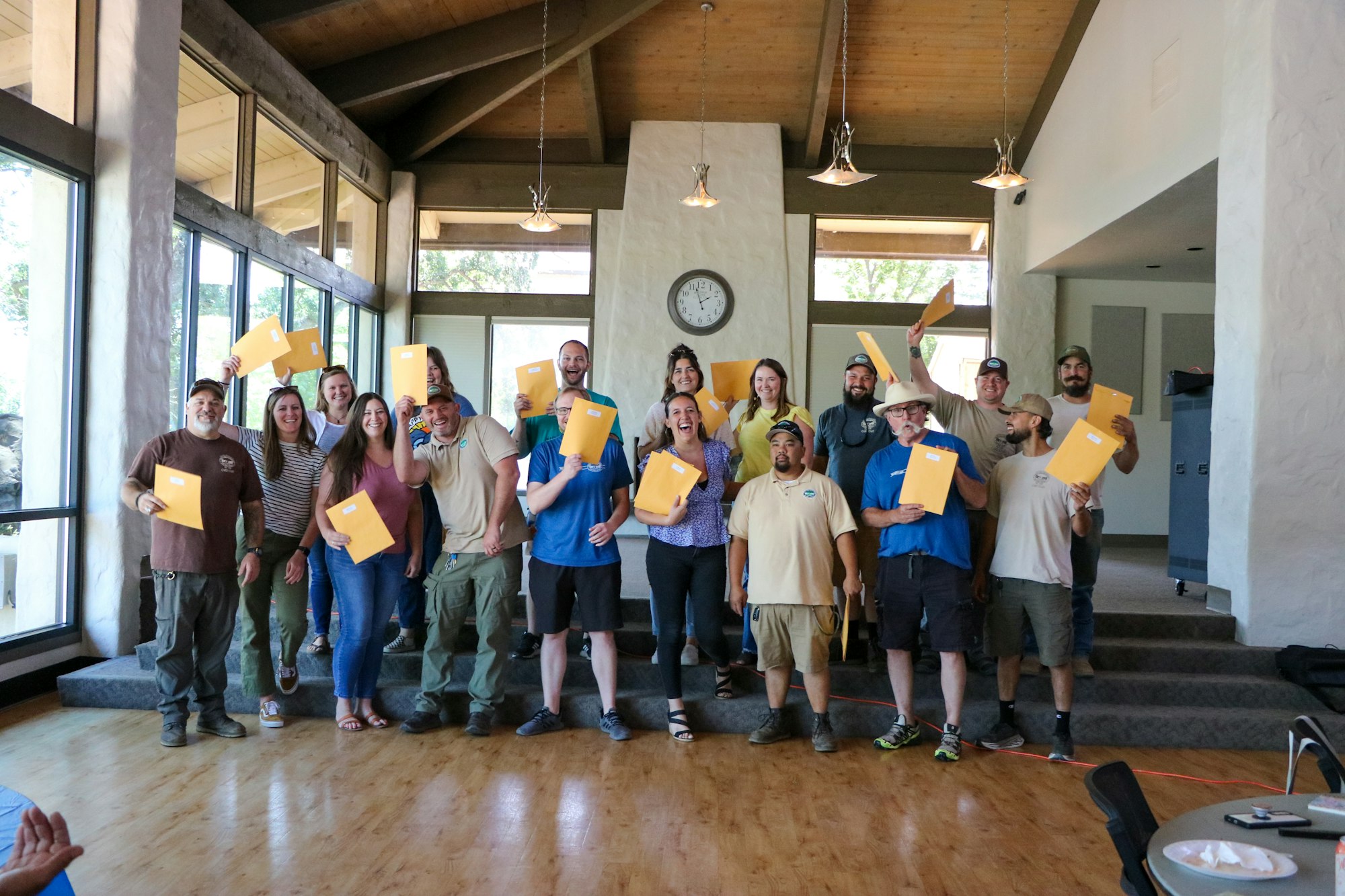A group of happy people holding envelopes, possibly celebrating an accomplishment or receiving awards, in a room with large windows and a clock.