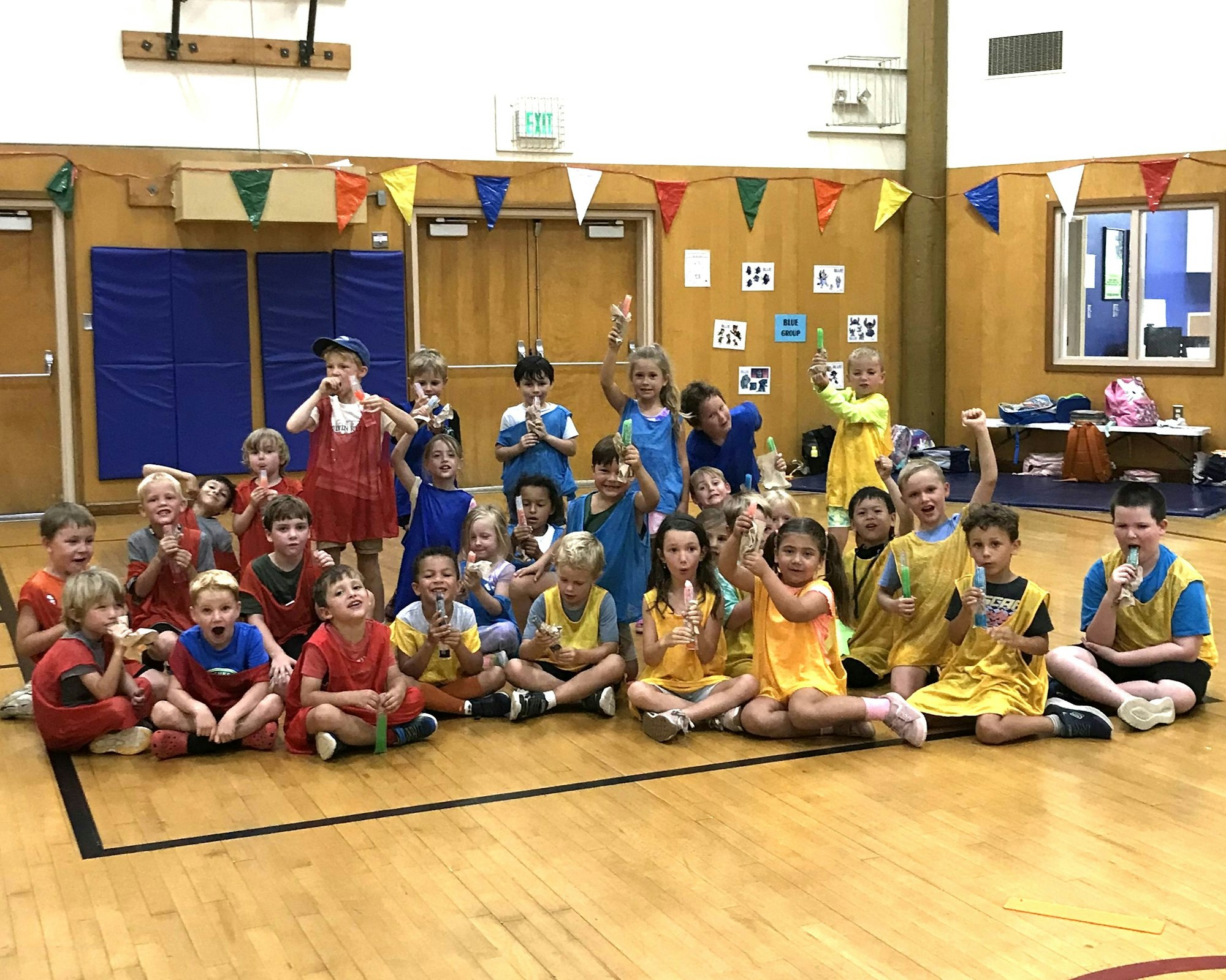 A group of children in colorful jerseys are joyfully posing with frozen treats in a gym setting.