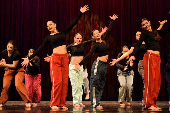 A group of dancers in crop tops and wide pants performing on stage against a colorful backdrop. Energy and expression are prominent.