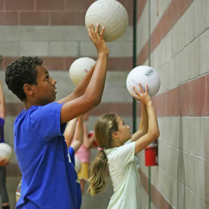 Kids are practicing with volleyballs against a wall in a gym.