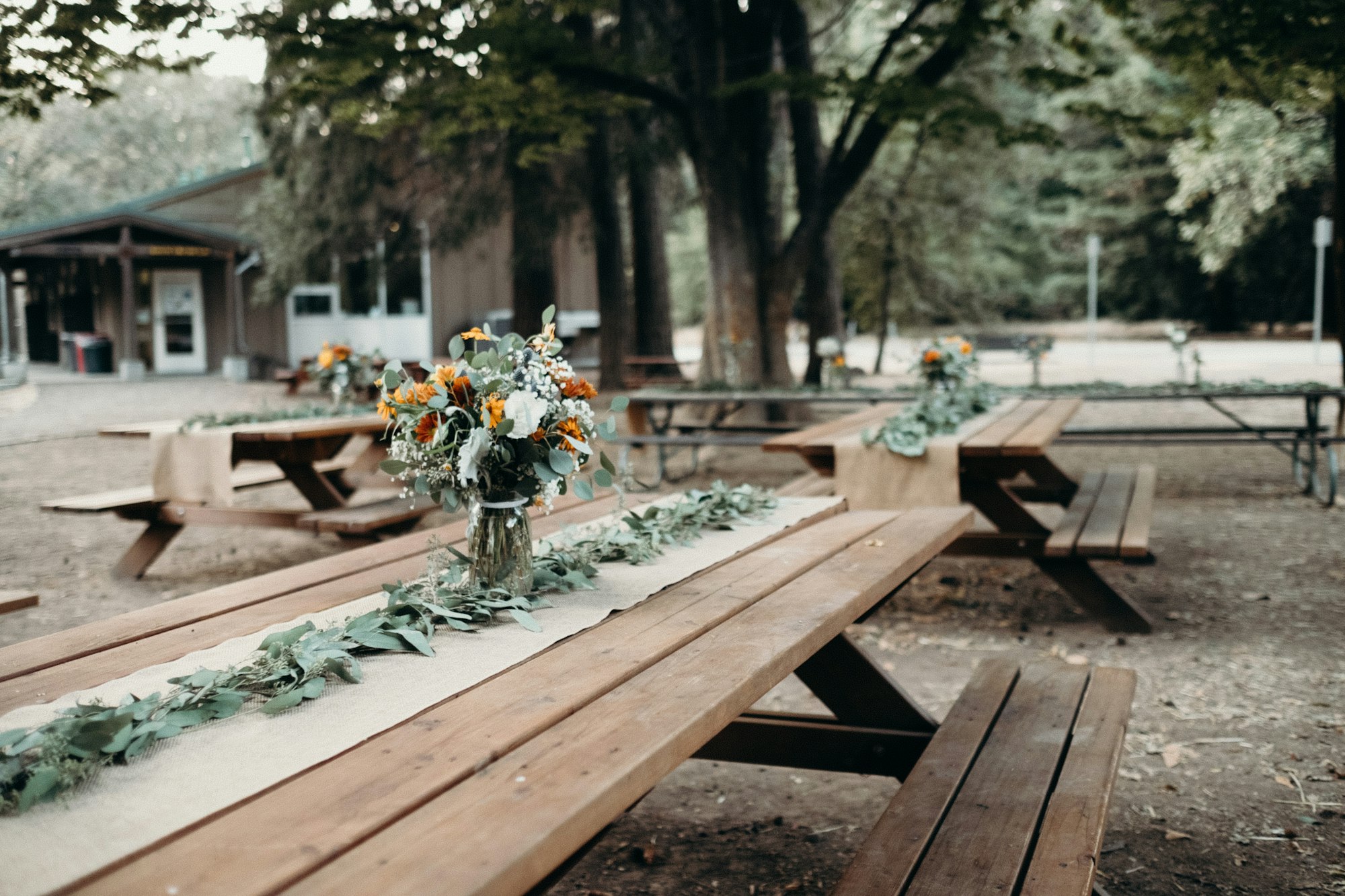 Outdoor setting with wooden picnic tables adorned with floral arrangements and greenery.