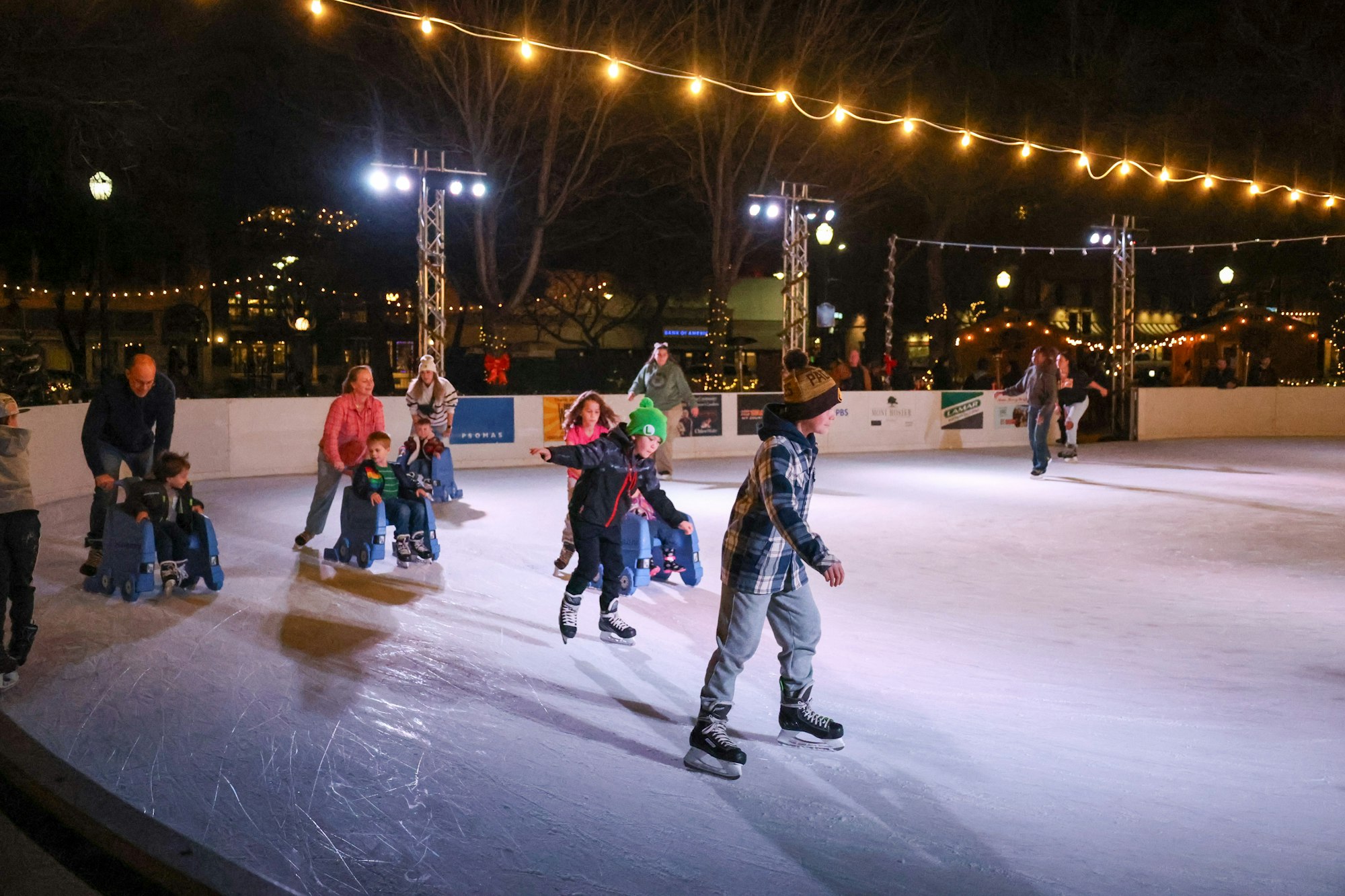 People ice skating on an outdoor rink at night, with string lights above and children in sleds.