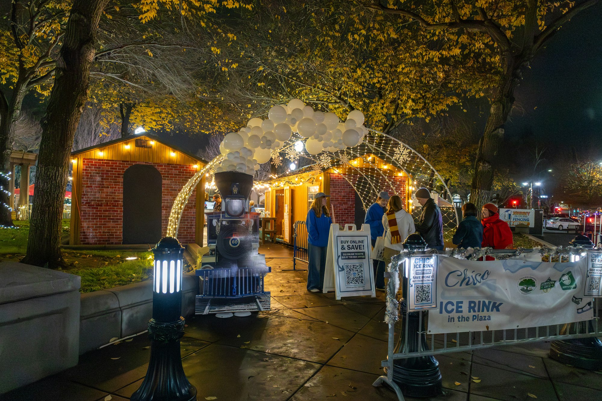 A festive outdoor scene at night featuring a decorated ice rink entrance, a mock train, and people gathering under lights.