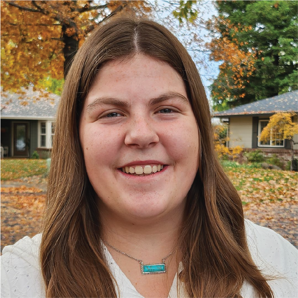 A person smiling outdoors in an autumn setting with houses and colorful foliage in the background.