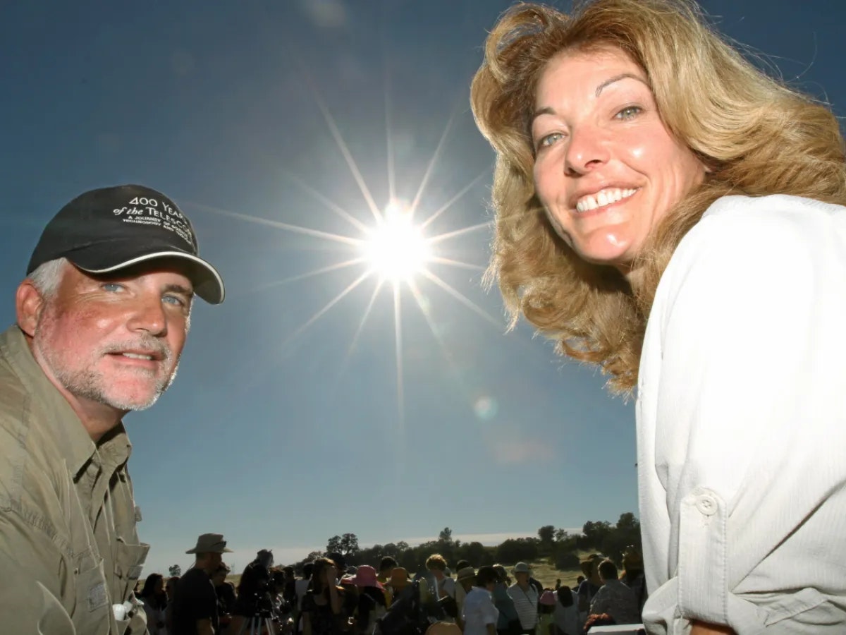 Two people smile outdoors under a bright sun. A crowd is in the background.