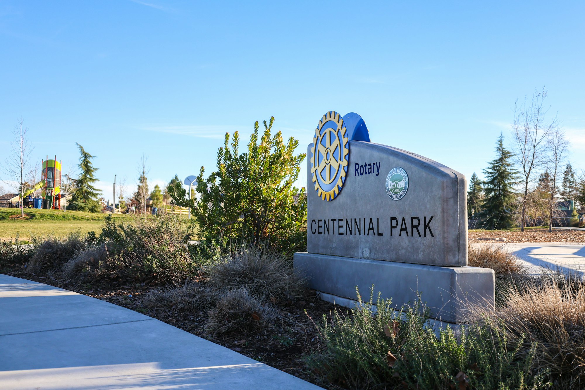 Centennial Park entrance sign with Rotary logo, surrounded by greenery and a playground visible in the background.