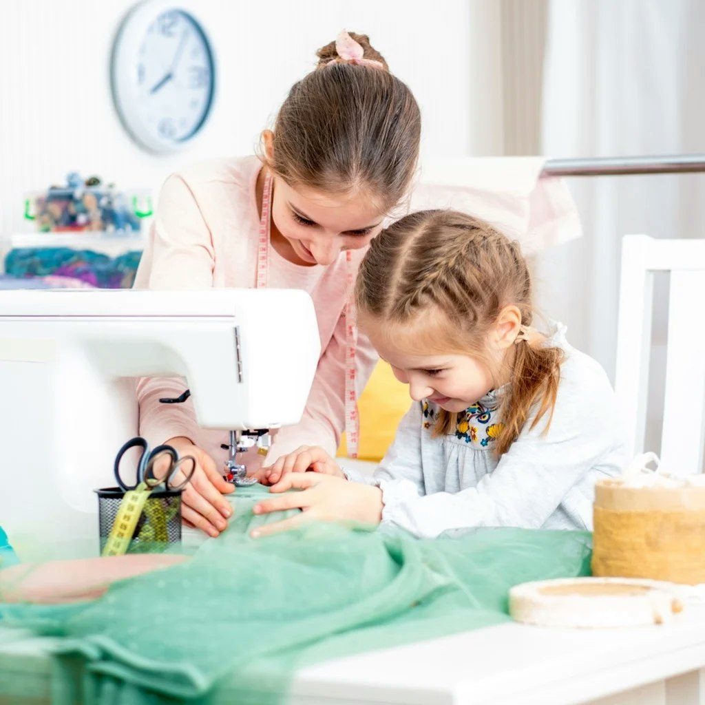 Two girls are working together at a sewing machine, focused on a green fabric, surrounded by sewing tools.
