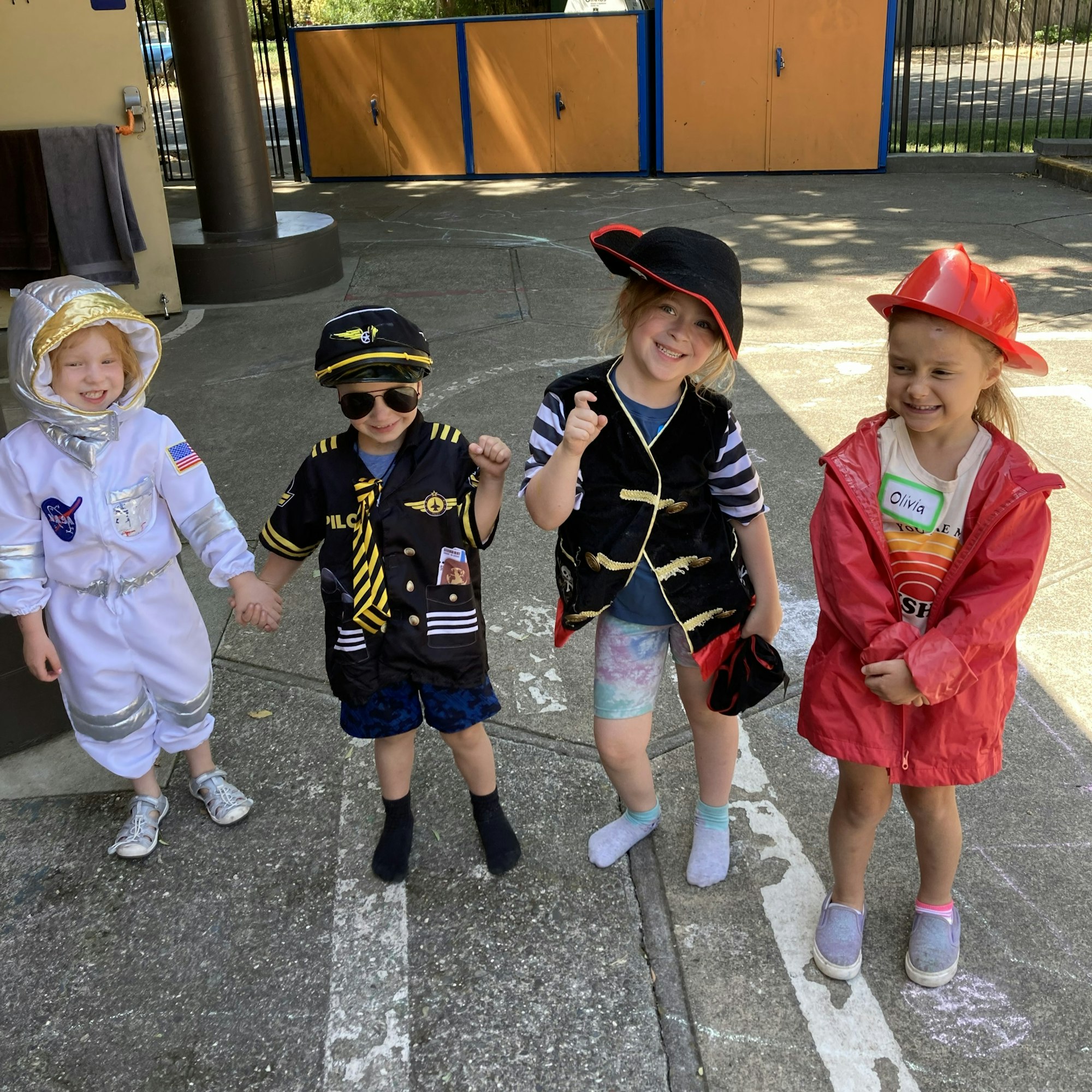 Four children in fun costumes: an astronaut, a pilot, a pirate, and a firefighter, smiling and posing outdoors.