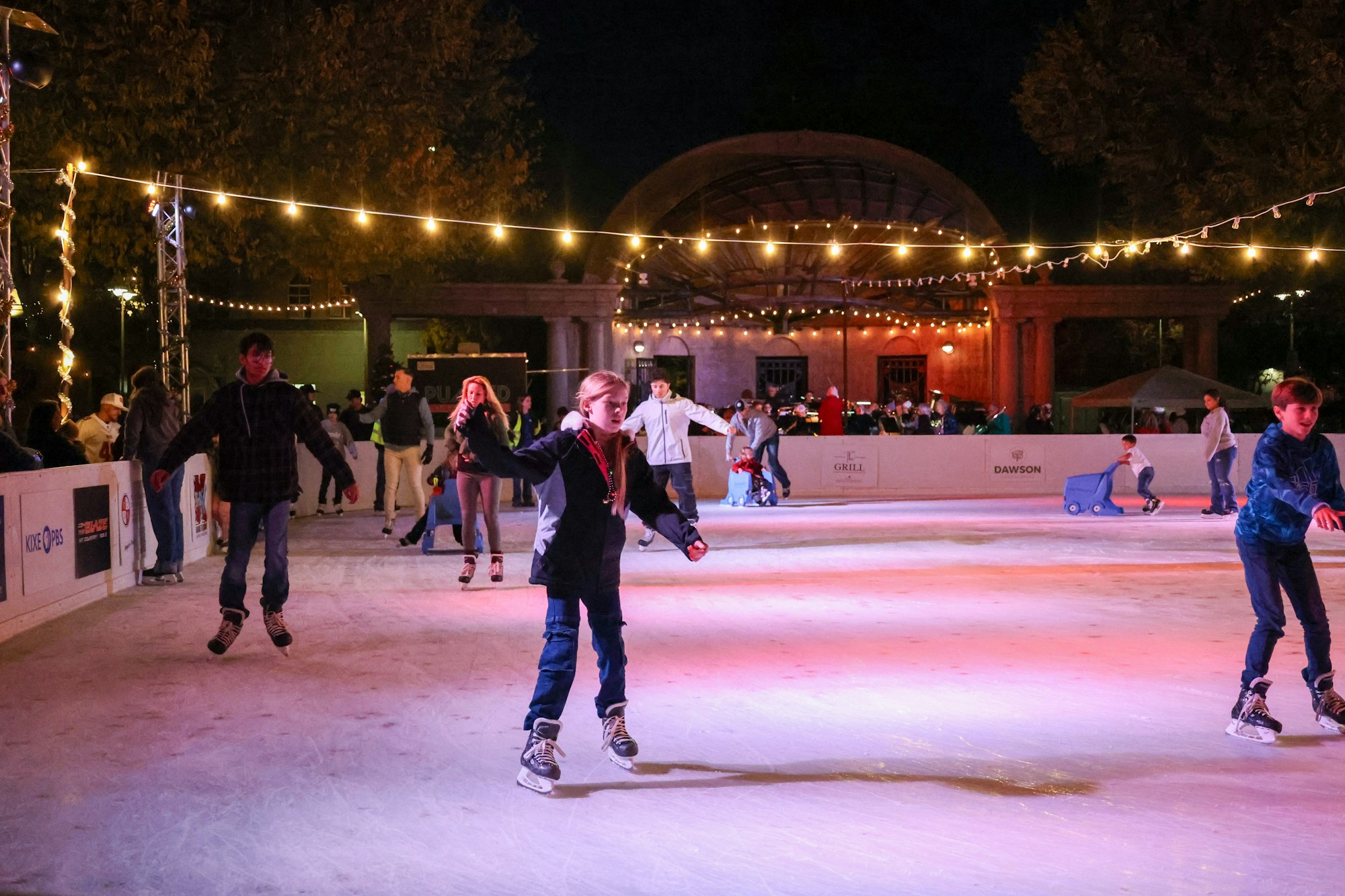People ice skating outdoors at night with festive lights overhead.