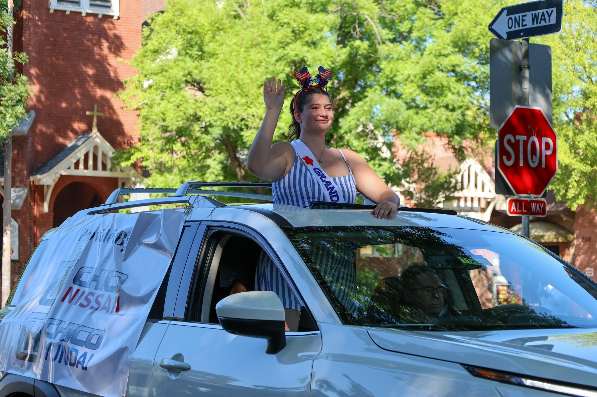 A person with a sash and headband waves from a car, part of a parade. Vehicle has a banner. Stop sign and church in the background.
