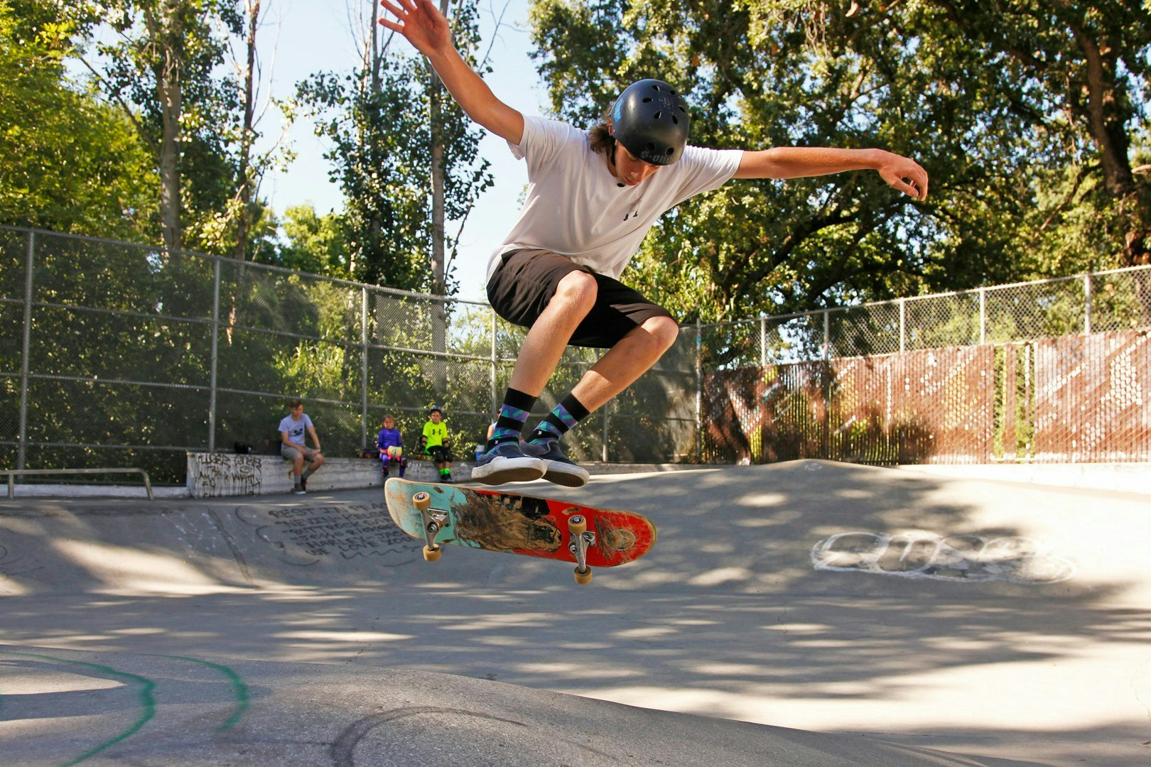 A skateboarder performing an aerial trick in a park, with spectators watching in the background.