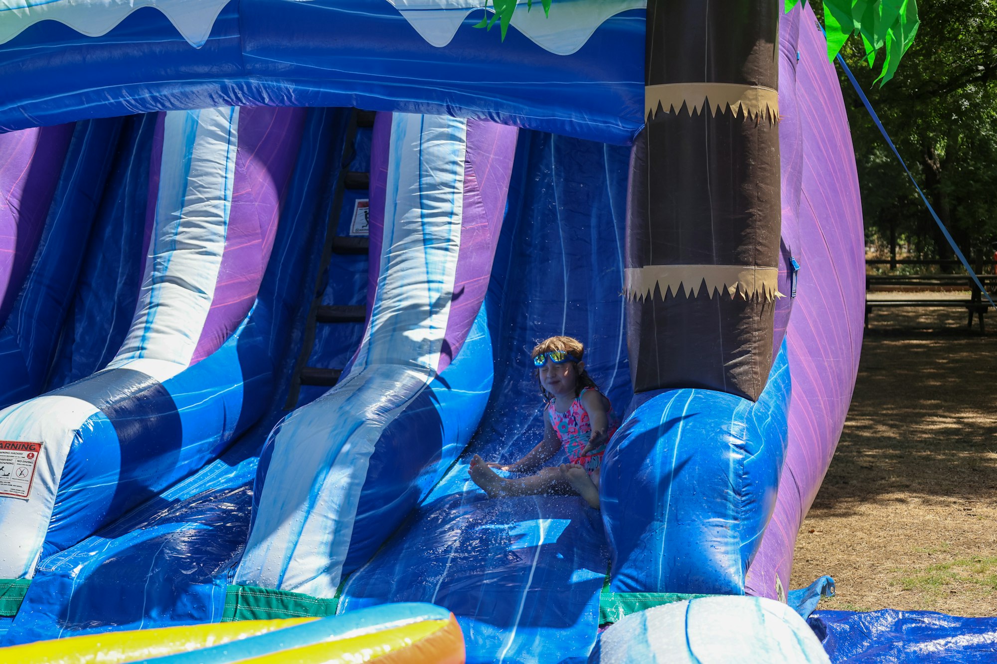 A child in goggles is sliding down a colorful inflatable water slide outside.