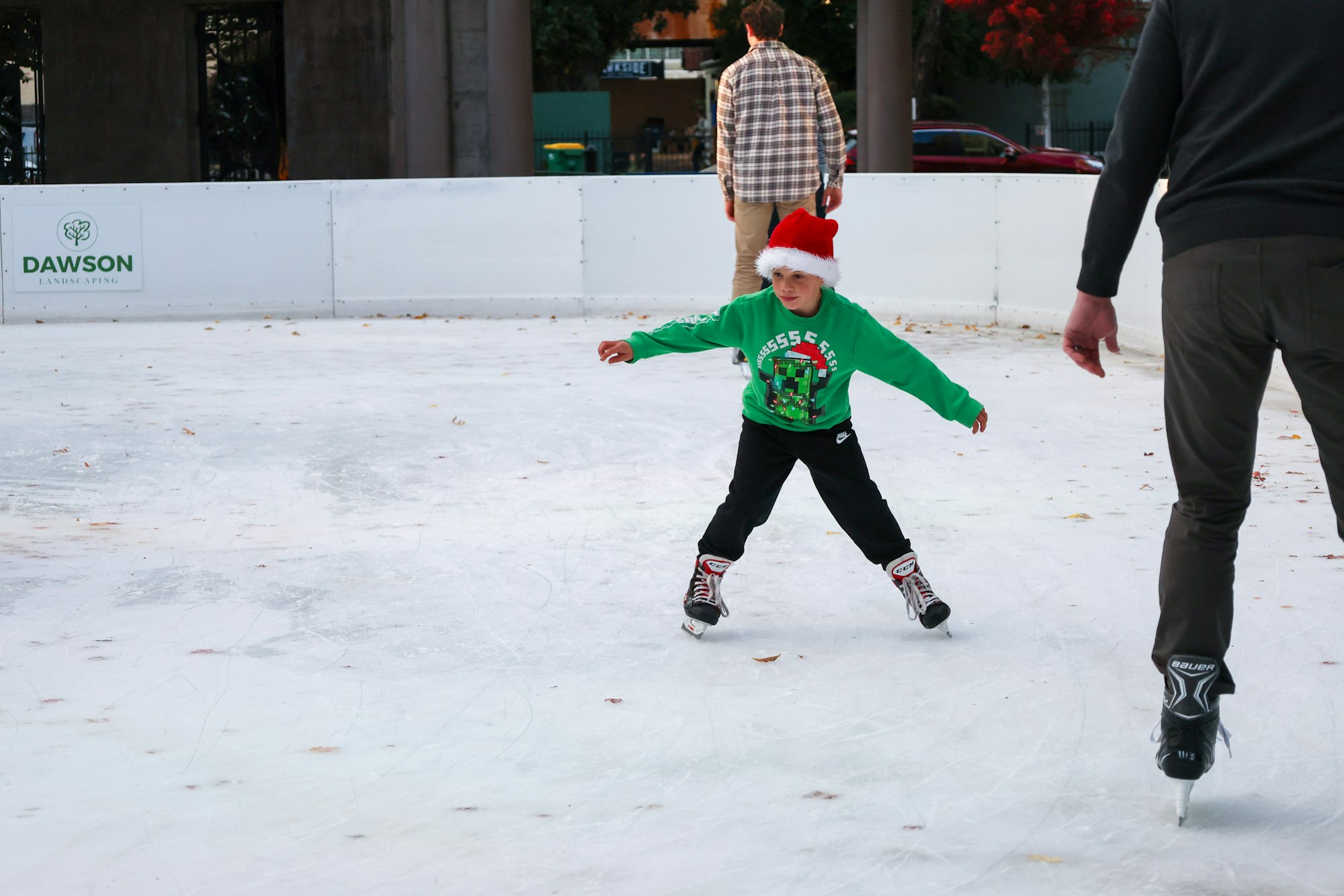 Child in green sweater and Santa hat ice skating with two adults nearby.