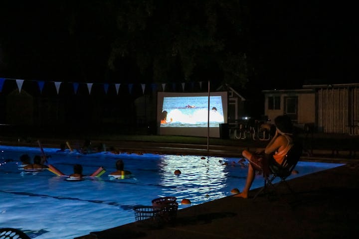 A nighttime poolside movie with people floating in the pool, watching a screen showing a film.