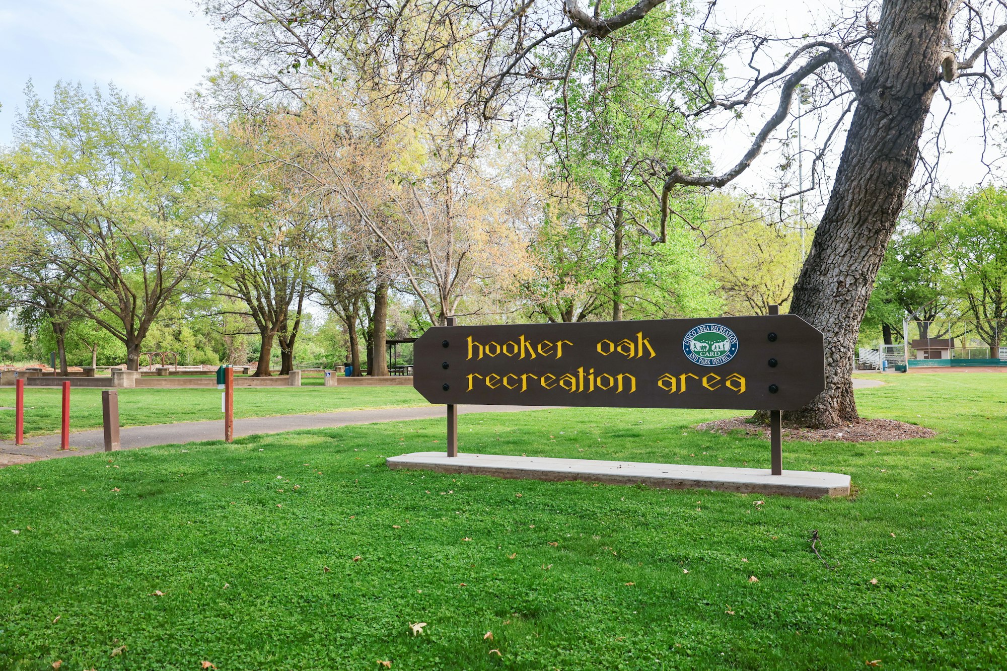 A sign for "Hooker Oak Recreation Area" in a lush park with trees, grass, and picnic benches.