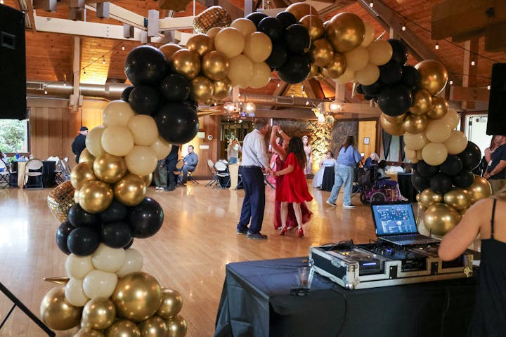 A festive venue decorated with black, gold, and white balloons, featuring people dancing and a DJ setup.