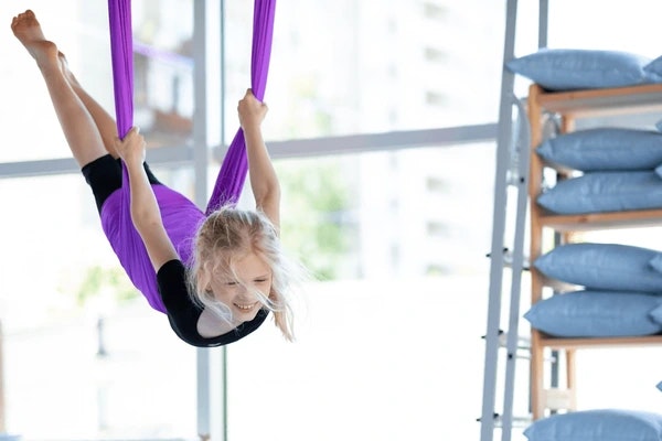A girl is happily practicing aerial yoga in a bright room, hanging upside down from a purple silk hammock.